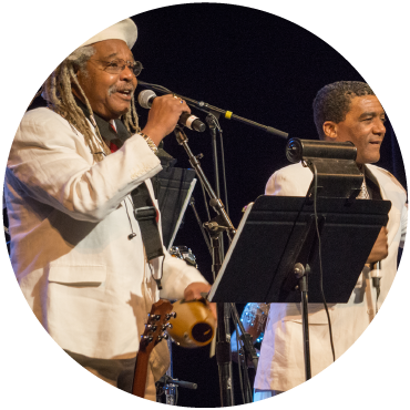 Juan de Marcos dressed in a white suite sings beside another performer in a white suit.