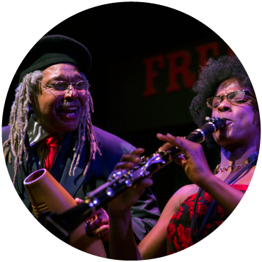 A performer in a dress plays the clarinet as Juan de Marcos looks on to them.