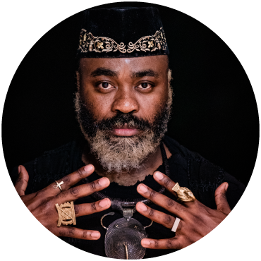 Nduduzo shows off the four rings on their hands in front of a black background. Nduduzo also wears a metal necklace and hat with gold stitching.