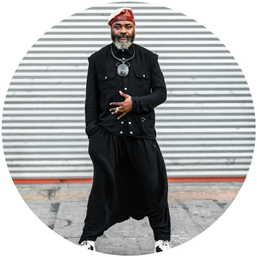 Nduduzo stands in front a gray and white horizontally-lined background and wears all black and a red hat.