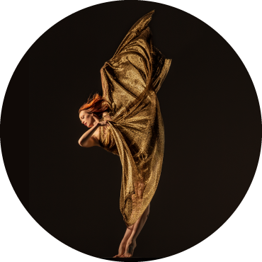 Dancer wears gold clothing and is captured in motion with her leg in the air in front of a black backdrop