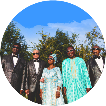 A circular image of the Blind Boys of Alabama, with Amadou and Mariam standing between them. The Blind Boys are wearing black suits with white shirts and bow ties. Miriam is wearing a white dress with a light green pattern and a light green head wrap. Amadou is wearing an aqua tunic with gold embroidery. They are dark skinned and wear glasses. Behind them are green trees and a blue sky with a white cloud. 