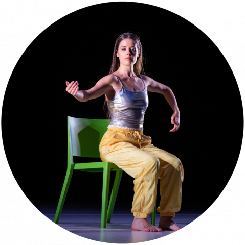 Jessie sits on a green chair wearing yellow pants and a silver tank top. Her legs are bent at the knee and her right arm is outstretched. Her left arm is bent down at the elbow.