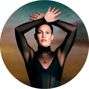 Carminho wears black and poses with her hands above her head against a sunset gradient backdrop