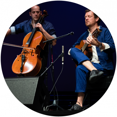 Derek Gripper wears a navy suit with a white button up shirt showing under the unbuttoned blazer. He holds his brown guitar and his eyes are closed. Mike Block stands to Derek's left holding his brown cello, wearing a chambray shirt with the sleeves rolled up