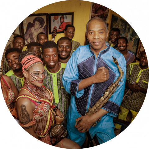 Femi Kuti stands in front of a group of other artists. He wears a blue outfit. His right hand is crossed over his chest, with his hand in a fist over his heart.