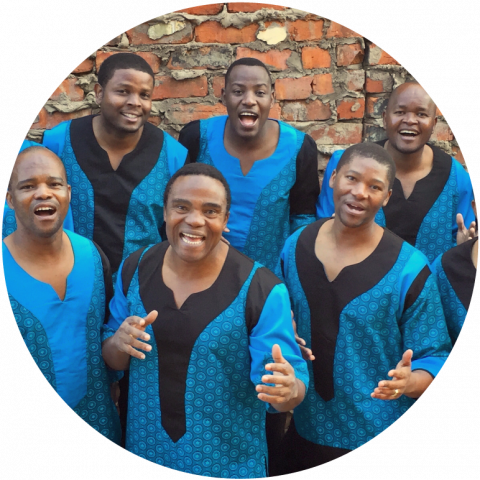 6 members look at the camera and stand in front of a red brick wall. They wear blue and black robes. 