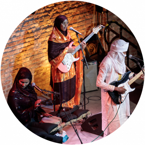 3 members of Les Filles de Illighadad are on stage i front of a brick wall wearing robes and head scarves. Two play guitars and the other plays the drums.