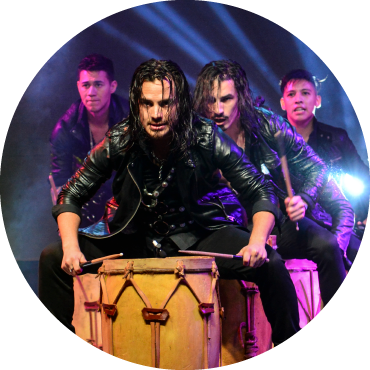 Four people stand crouched in front of large drums in front of them, all with drum sticks in their hands. The two people in front have long black hair, while the others have short black hair. They all wear black pants and leather jackets.