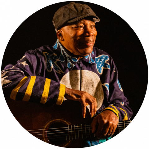 Milton wears a black cape and a zip up blue jacket with yellow strips on the sleeves and a big white circle on the chest. He holds his guitar across his lap with a smile on his face.
