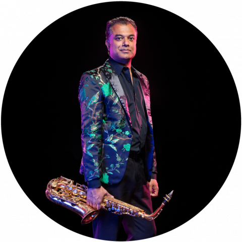 Rudresh stands in front of a black background. He has dark skin and wears a black tie-up shirt and pants, with a jacket with a flower pattern. He looks at the camera and holds a saxophone at his side.