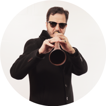 Mehmet Ali Sanlıkol wears a black suit with sunglasses and poses against a white backdrop, playing the Zurna
