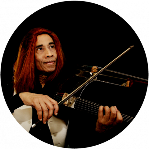 L. Shankar is front of a black background and plays a 10-string double violin. He has dark skin and red hair that goes past his shoulders.