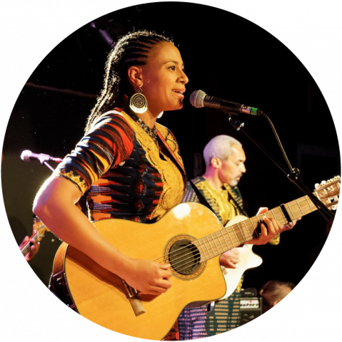 Sona is on stage singing into a microphone and plays an acoustic guitar. She has dark skin and hair that is breaded and wears a colorful shirt and gold earrings.