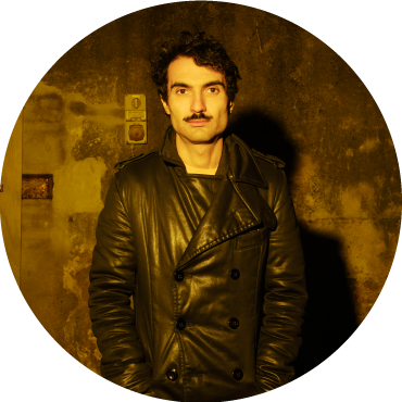 Tigran Hamasyan stands in front of a textured concrete wall lit by yellow lighting.