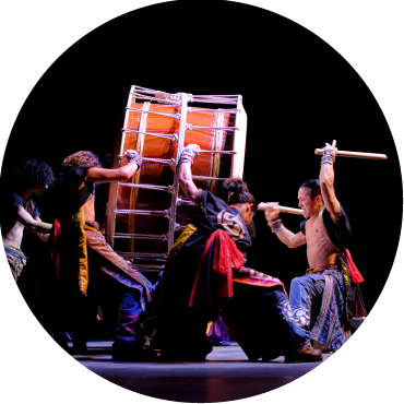 A circular image of a group of men with long black and brown hair are dressed in red and black. They are holding up a large drum which is being played by a man in blue pants and an open black shirt. 