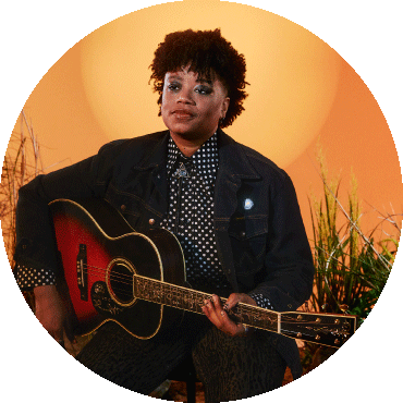 Amythyst Kiah holds a guitar and poses by a sunset orange background,