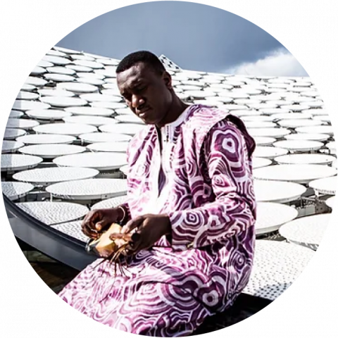 Bassekou wears a purple and white traditional African shirt holds his ngoni sitting on the edge of a white art installation