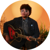 Amythyst Kiah poses with a guitar against a sunset orange background,