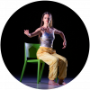 Jessie sits on a green chair wearing yellow pants and a silver tank top. Her legs are bent at the knee and her right arm is outstretched. Her left arm is bent down at the elbow.