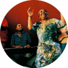 Cesaria Evora is smiling with their mouth open and their hands are raised. They are wearing a green and yellow patterned dress and are standing in front of a band.