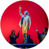 Farruquito stands on a platform surrounded by other performers. He wears a white suit and faces to the right with his right hand raised above his head 