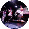 Red Baraat band members are on stage holding various instruments and are all wearing white jumpsuits with flowers painted on. The two members in the center are jumping.