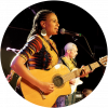 Sona is on stage singing into a microphone and plays an acoustic guitar. She has dark skin and hair that is breaded and wears a colorful shirt and gold earrings.