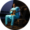 Vieux Farka sits with a guitar by a shore at night.