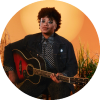 Amythyst Kiah holds a guitar and poses by a sunset orange background,