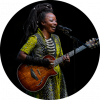 Singer Natu Camara wears a yellow, black, and white-dotted dress as she a brown guitar and sings into a microphone.