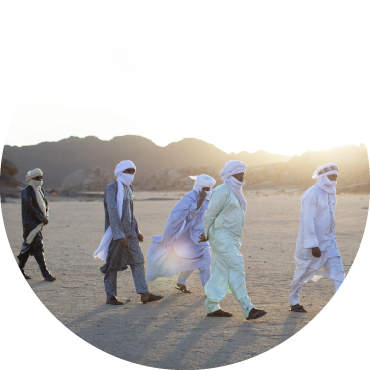 6 members of the band wearing full scarfs walk through the desert with the sun behind them